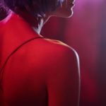 A woman in profile with a purple wig, illuminated by neon light, wearing a spaghetti strap dress.